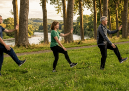 Zwei Männer, eine Frau führen Taiji im Park aus. Neben der Wiese stehen Bäume, durch ihre Stämme sieht man einen Fluß
